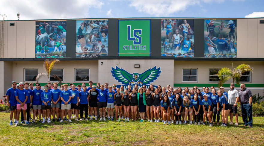 LSSC student-athletes pose in front of a building displaying the LS Athletics logo and a hawk mascot mural.