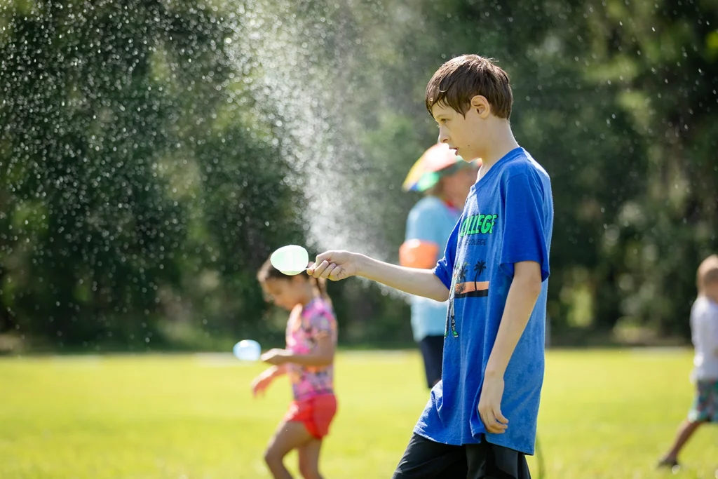 Person in blue shirt holding water balloon outdoors, with others playing and water spraying in background.