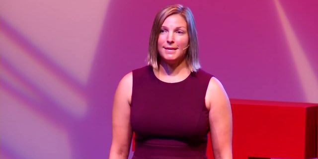 Women in burgundy dress speaking on stage with purple backdrop