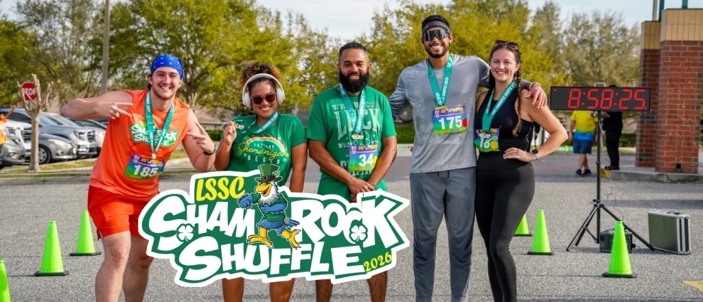 Five smiling race participants pose with medals at the finish line of the Shamrock Shuffle 2026, hosted by the Lake Sumter State College Foundation. A race clock shows 58:25, with trees, cars, and a brick building in the background.