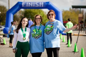 Three individuals at race finish line; two in blue Shamrock Shuffle shirts, one displays race medal; outdoor event setting.
