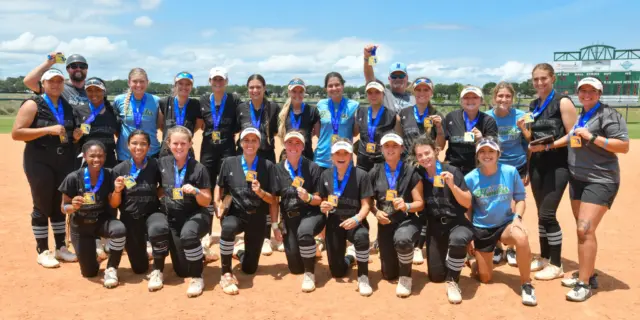Softball team in black and blue uniforms on dirt field, each person holding a gold medal.