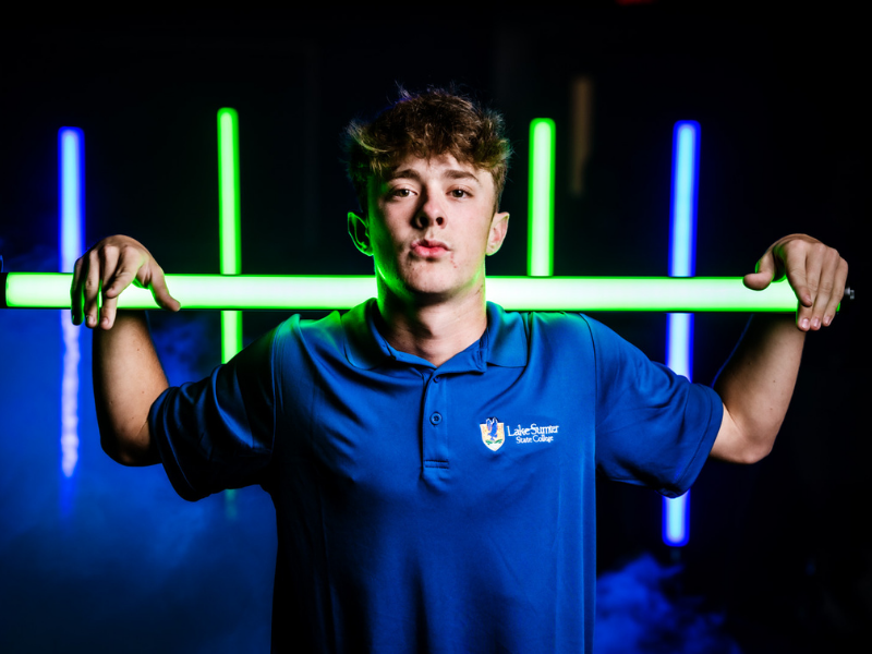 Person holding a glowing green bar across shoulders while wearing a blue Lake‑Sumter State College polo shirt, standing against a dark background with vertical neon green and blue light bars and light fog.