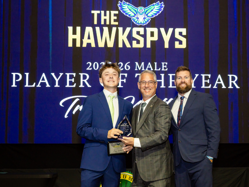 Three individuals on a stage holding a trophy in front of a large screen that reads “THE HAWKSPYS 2025–26 MALE PLAYER OF THE YEAR.” One individual in a blue suit accepts the award while two others stand beside them, with a Lake‑Sumter State College emblem visible below the screen.