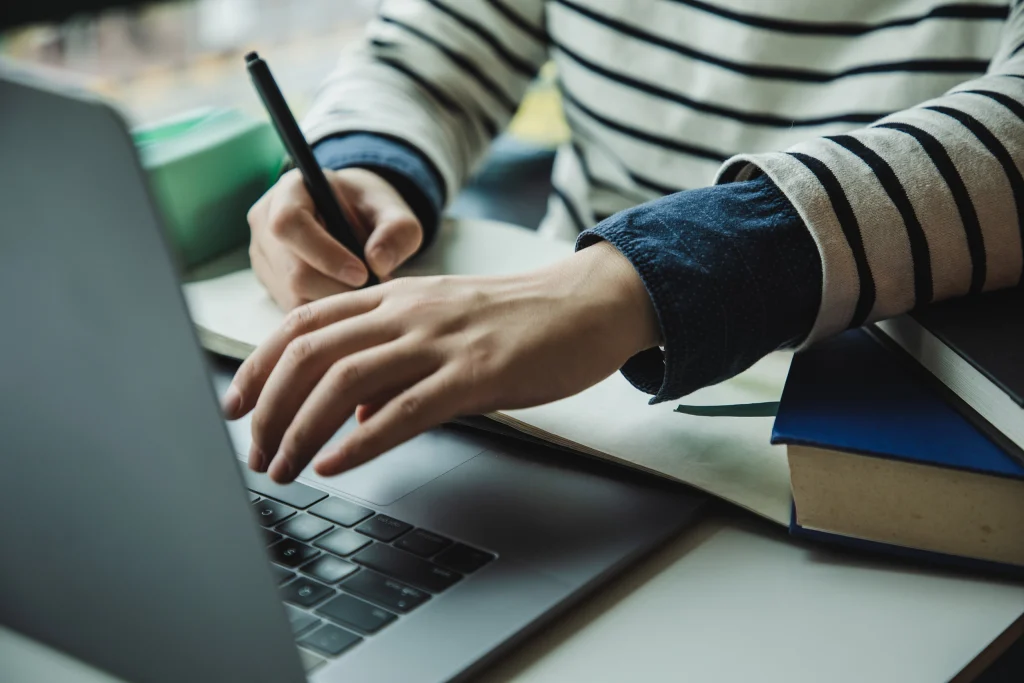 Student with striped shirt writing in notebook, using laptop, books stacked on desk; study setting.