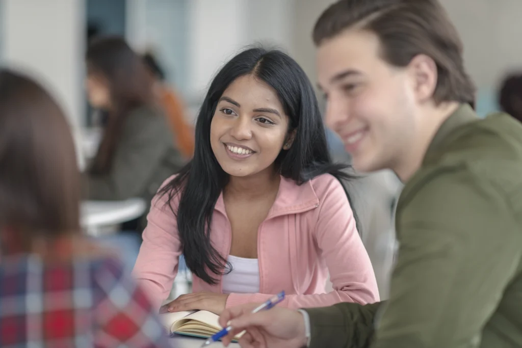 Students at a table smiling, talking, and taking notes with others in a bright classroom.