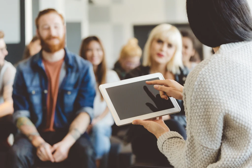 Person with tablet gesturing, group listening attentively in casual setting (person teaching).