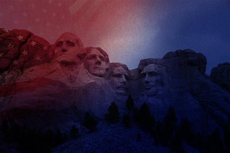 Mount Rushmore at dusk with American flag overlay, darkened sky. Monument and flag visible, evening setting.