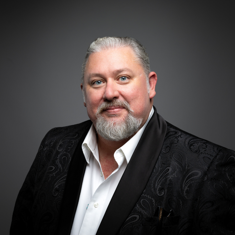 A man with short hair and beard wears a black patterned jacket over a white dress shirt, posing against a gray background.
