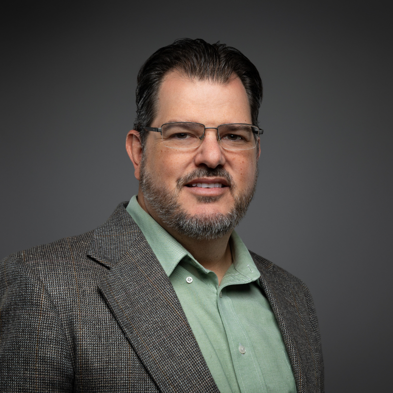 A man with dark hair, glasses, and a beard wears a gray plaid blazer over a light green shirt, standing in front of a neutral gray background.