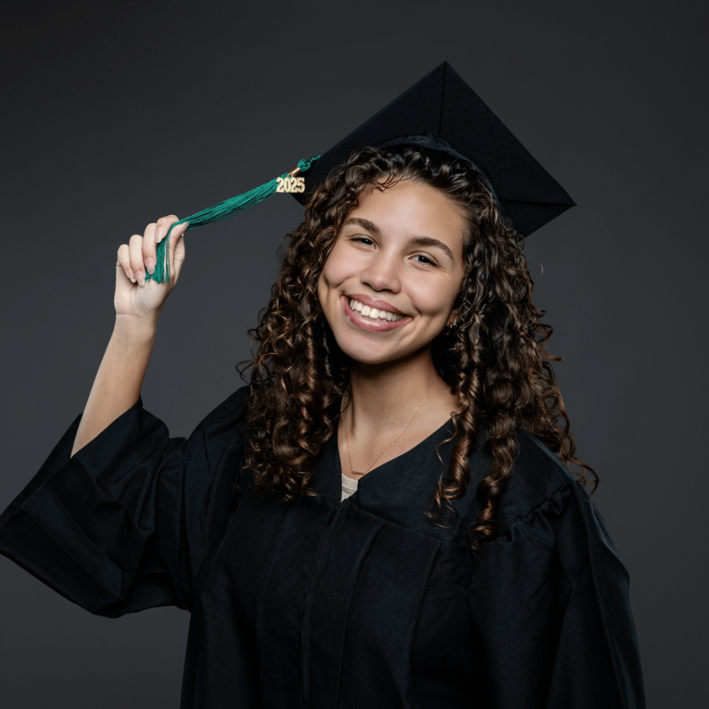 Graduate in a black cap and gown stands against a dark gray background, smiling and lifting the green “2025” tassel on the cap with one hand.