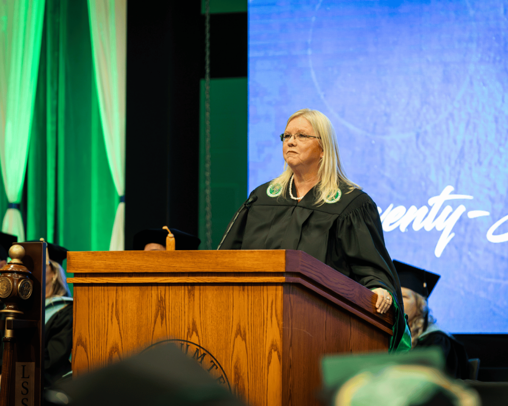 Woman in black academic regalia stands behind a wooden podium on a brightly lit commencement stage, speaking into a microphone