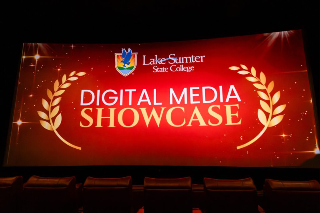 Group of individuals on stage holding awards at event, with plants and flags in background.