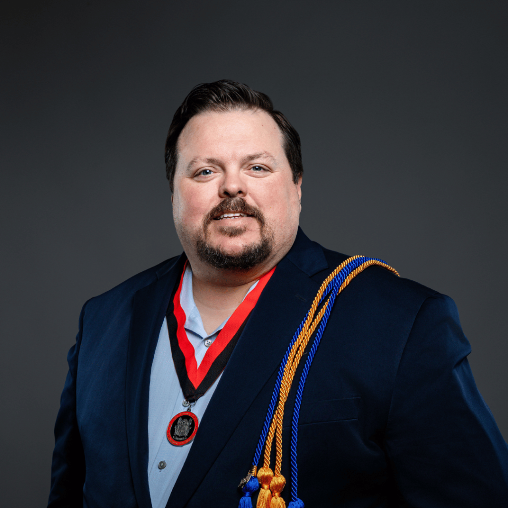 Graduate in a navy suit jacket and light dress shirt stands against a dark gray background, wearing multiple honor cords over one shoulder and a red and black medallion around the neck.