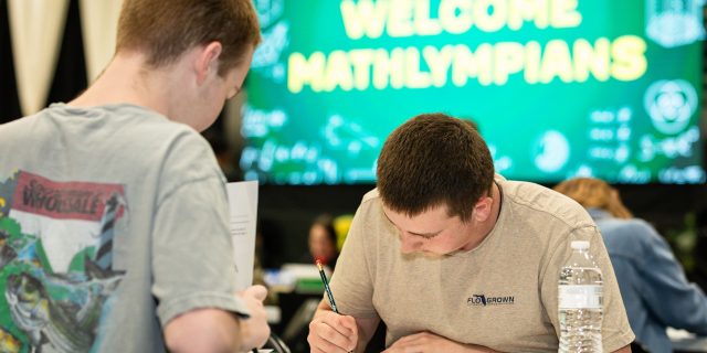 Two students with papers at table, large screen behind reads “Welcome Mathlympians” in bright yellow. Classroom setting.