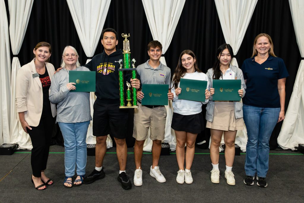 A group of seven people stands on a stage in front of black-and-white draped curtains, with several holding green diploma folders and one person holding a tall trophy.
