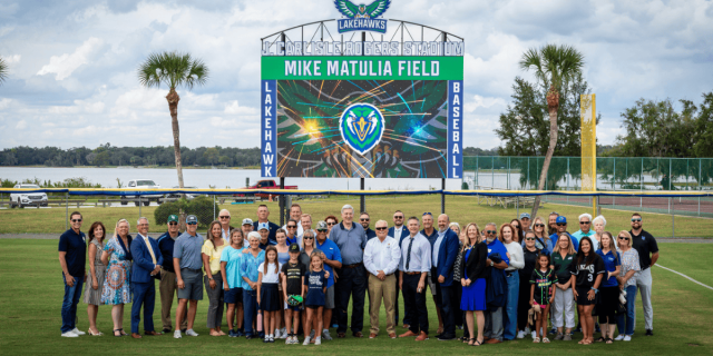 Group of people gathered on a baseball field in front of a scoreboard for the Lakehawks’ Mike Matulia Field dedication.