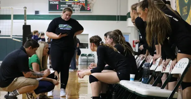 Individuals in volleyball uniforms gathered on the sidelines of a gymnasium.