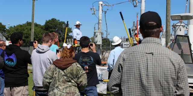 back of crowd outside looking at an electrical pole demonstration