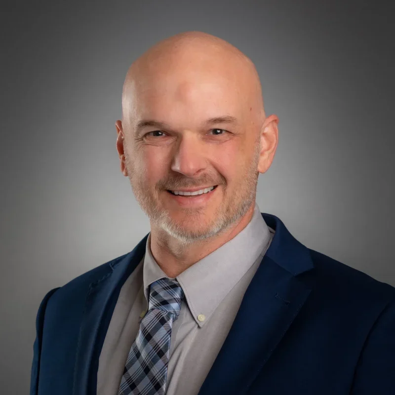 Jason Myer in blue suit, gray shirt, plaid tie, smiling; bald with beard; gray background. Setting undetermined.