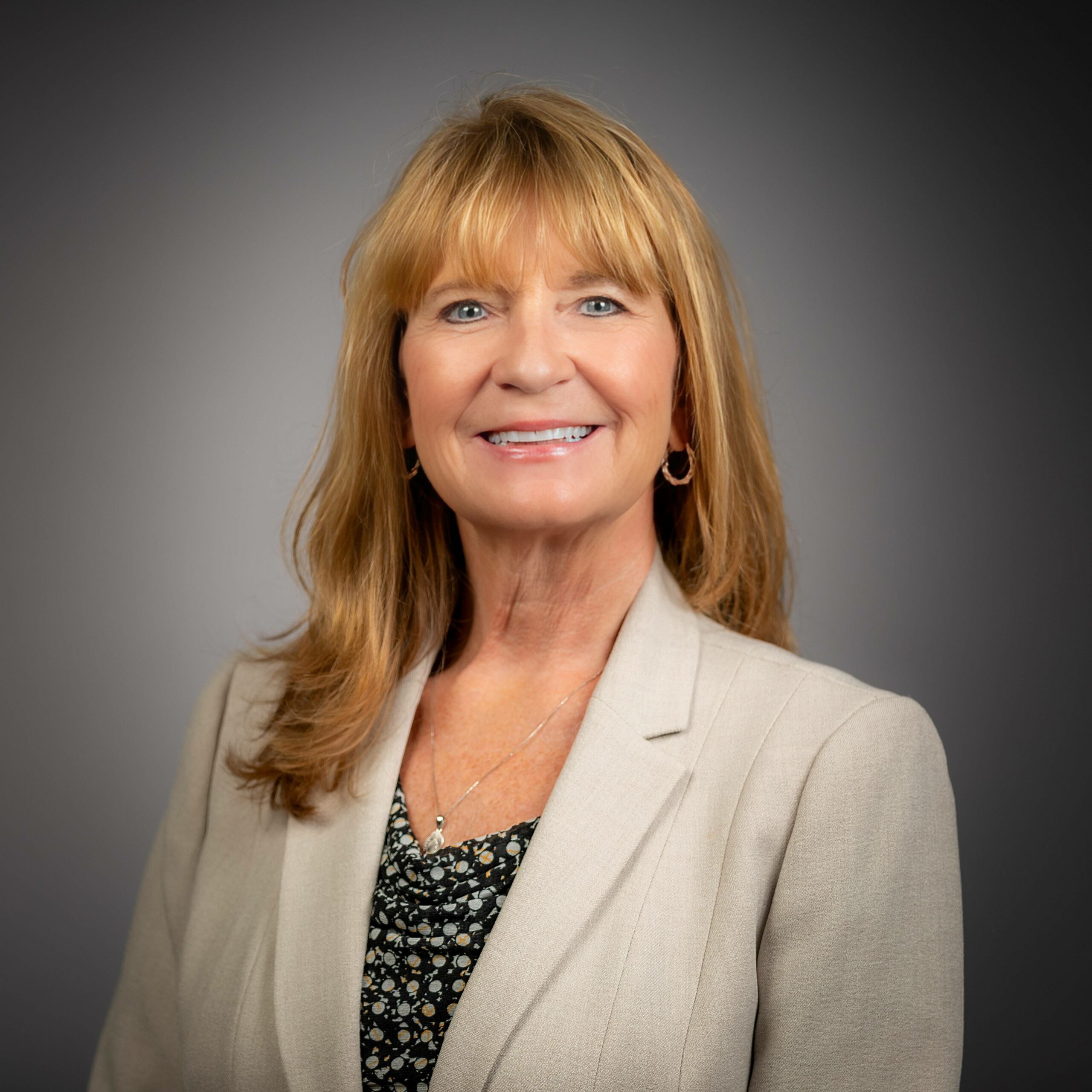 Smiling person with long blonde hair in beige blazer and patterned top, gray background.
