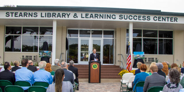 Man standing at a podium in front of a building speaking to a seated crowd