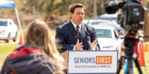 Florida Governor Ron DeSantis speaks at a podium with cars lined up for vaccinations behind him and news cameras facing him