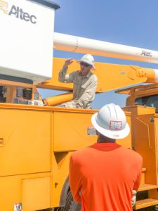 Man in hard hat sits on yellow utility truck explains something to student looking up at him