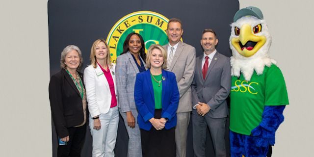 Six people in business attire pose for a photo with the school mascot