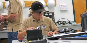 Man in a khaki colored shirt working on wiring while sitting at a table