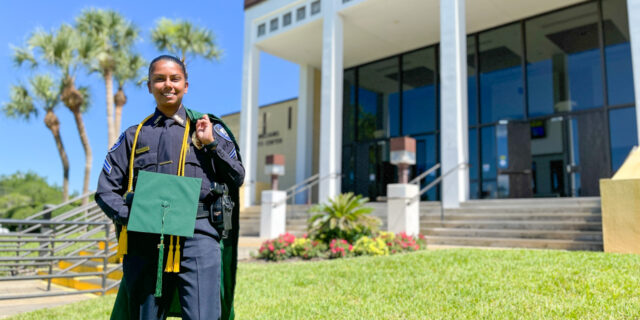 Female student in police uniform holding graduation cap and gown standing in front of campus building with palm trees and blue sky in background