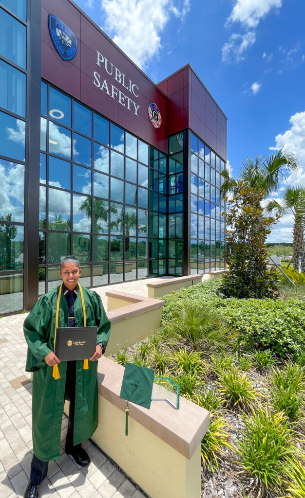 Female student in police uniform and green graduation regalia standing with diploma and awards in front of Public Safety building