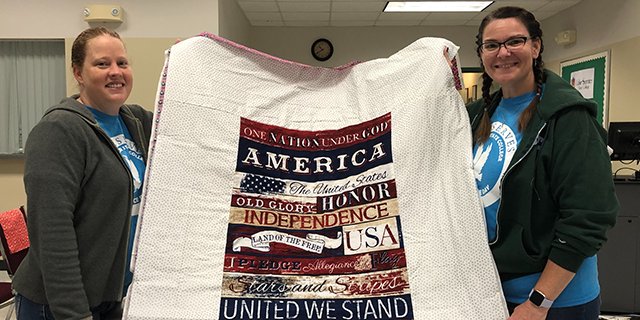 Two females stand holding a patriotic themed quilt