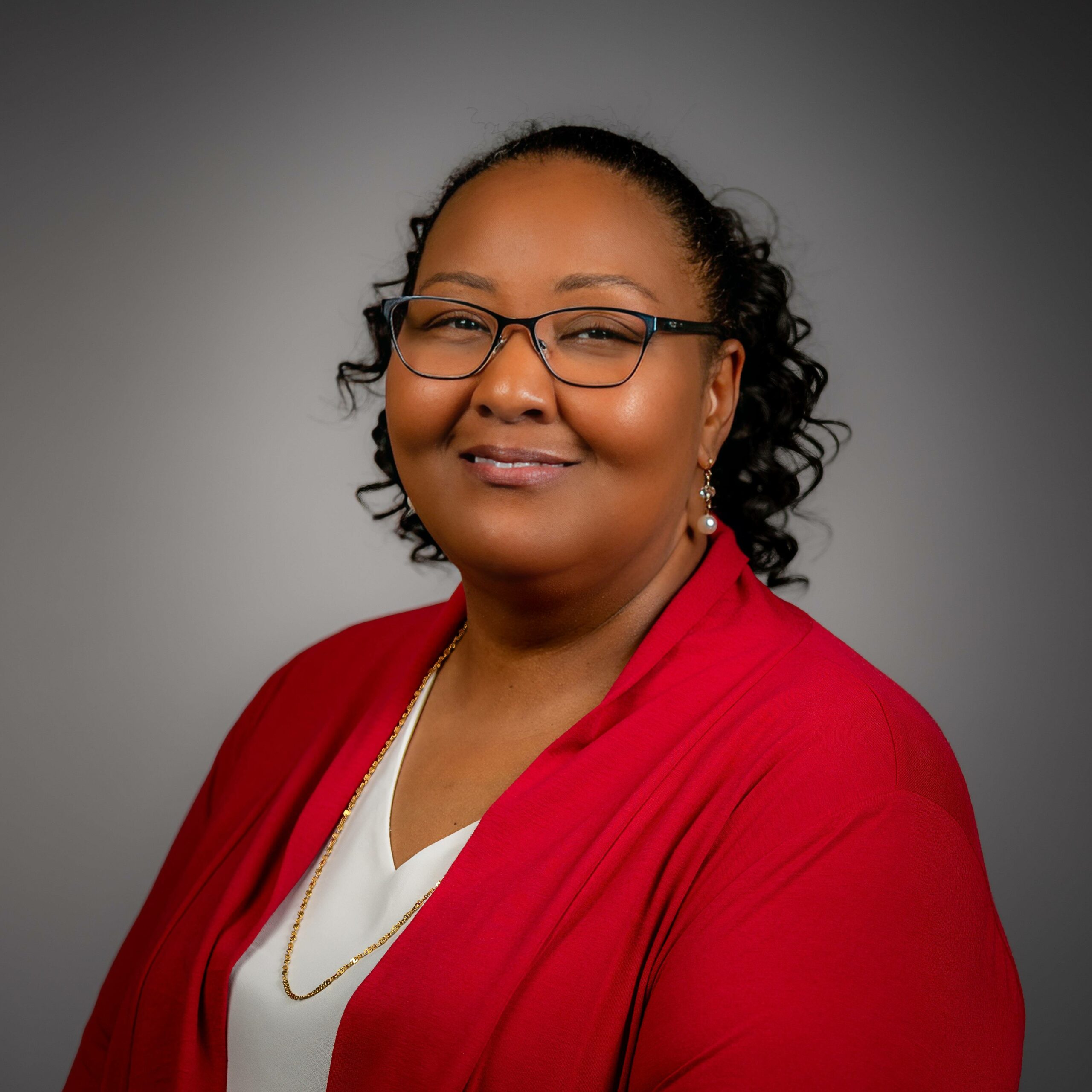 Person with glasses and curly hair in a red blazer and white blouse, smiling in front of a plain gray background.