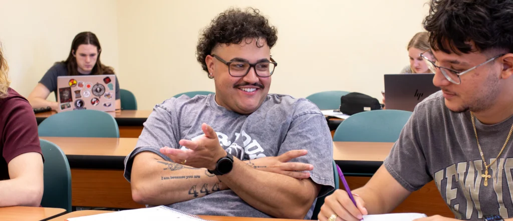 Students discussing course accessibility in classroom; others using laptops in background.