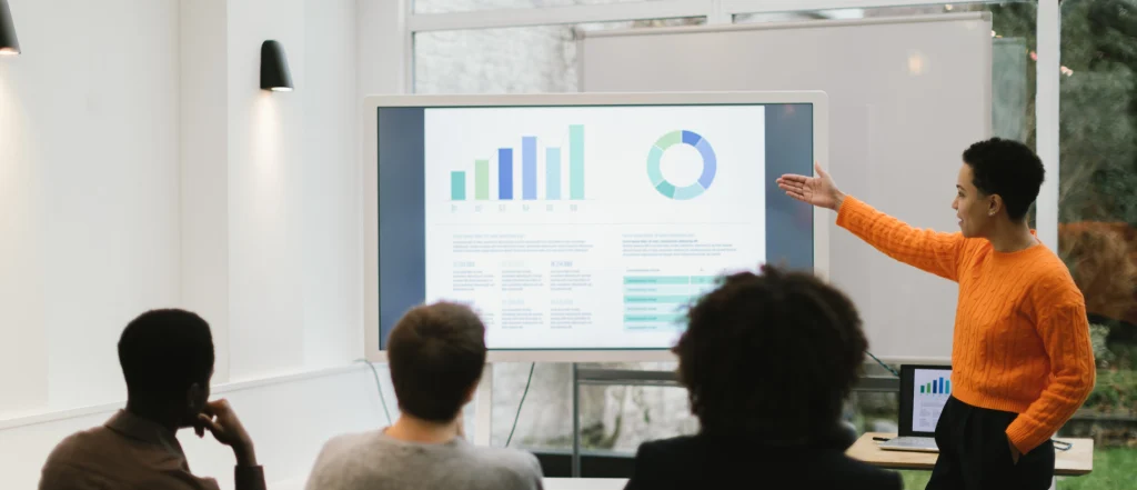 Person presents charts and graphs on large screen to audience in modern meeting room (person teaching).