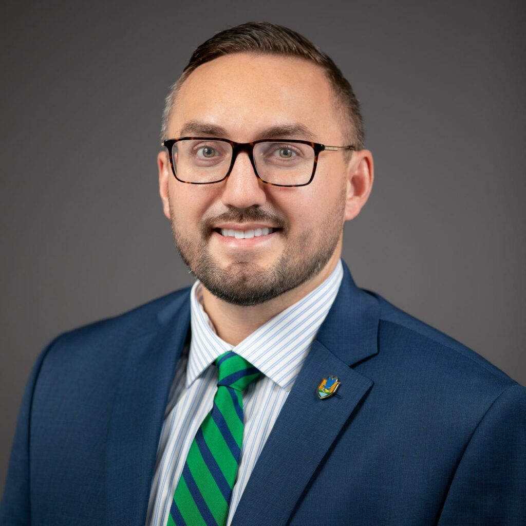 Portrait photo of Dr Joseph Mews in a blue suit with a green tie