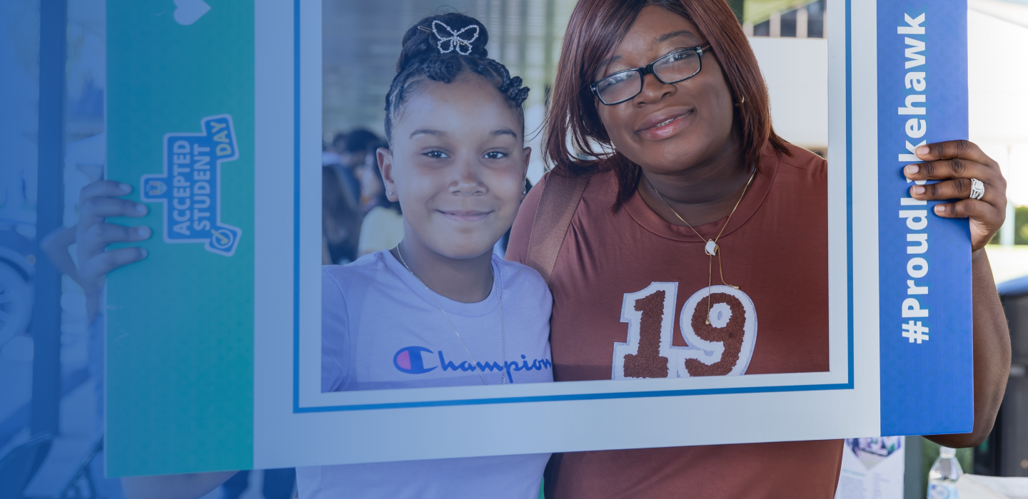 Two individuals smiling, holding large photo frame with #ProudLakehawk and Accepted Student Day text. Indoor, neutral setting.