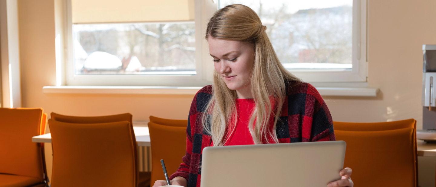 Staff at desk, registering for courses on laptop and writing in notebook in bright room with orange chairs.
