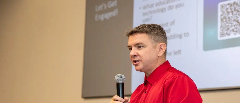 Person in red shirt speaks into microphone with projected slide behind, presenting in lecture hall.