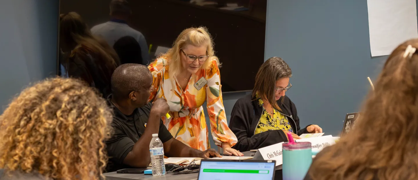 VP Tolx in floral dress assists staff at table during meeting or workshop; collaborative setting.