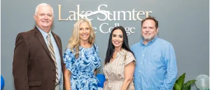 Four individuals smiling in front of a Lake-Sumter State College sign, wearing business and casual clothing.