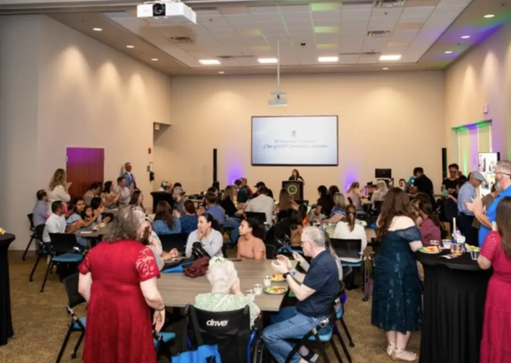 Individuals seated at round tables, mingling in modern event room; presentation screen visible at front.