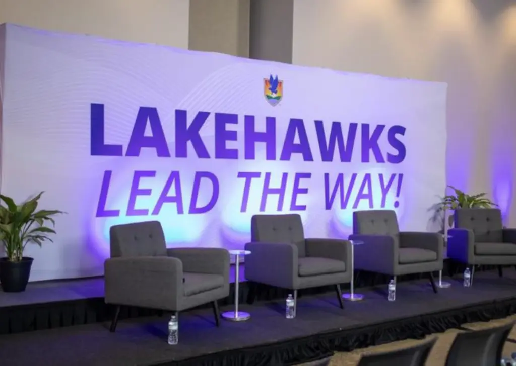 Stage in Cooper Memorial Libray with four gray chairs, small tables; LAKEHAWKS LEAD THE WAY! displayed on backdrop. No people present.
