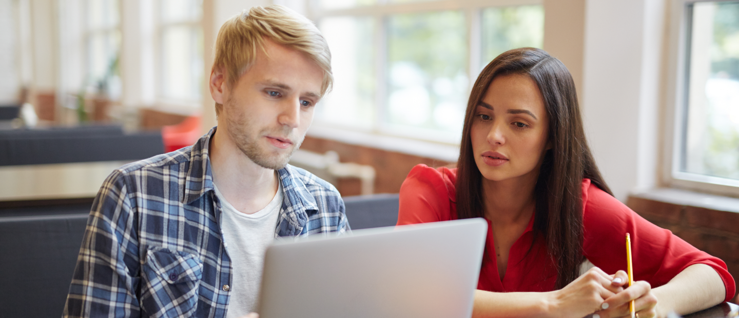 Two individuals discuss course registration while viewing a laptop, seated near bright windows. Setting unclear.