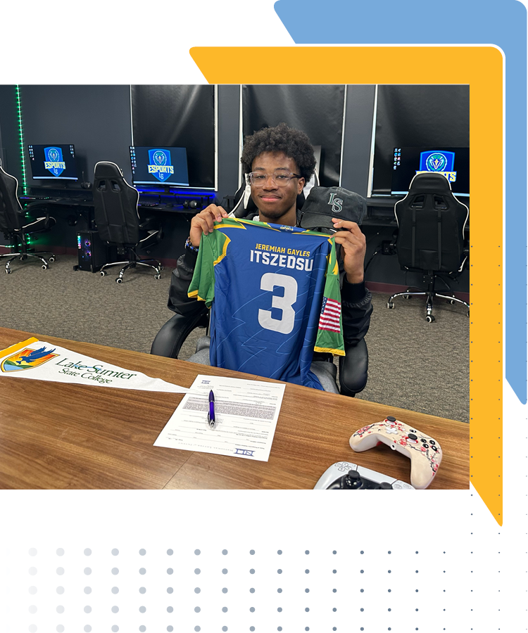 A person in a gaming room holds up a blue Lake-Sumter State College esports jersey with the name "Jeremiah Gayles," username "ITSZEDSU," and number 3. On the desk are a college pennant, a document with a pen, and two gaming controllers. Behind them are gaming chairs and monitors showing the esports logo
