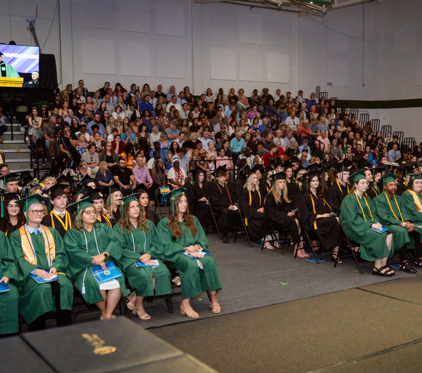 Students in green graduation attire seated in rows; audience in bleachers behind. Commencement ceremony setting.