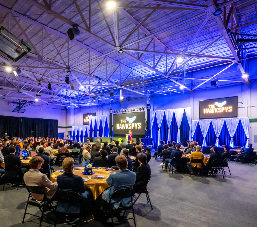 Individuals at round tables watch a person on stage at The Hawkspys indoor event with blue and white decor.