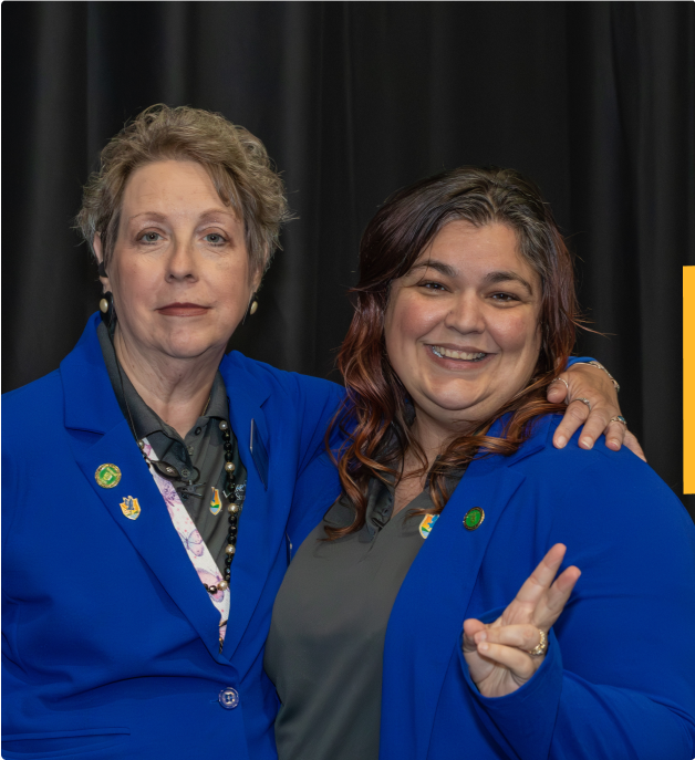 Julie Murray and Sherry Pelfrey in blue LSSC branded sweaters with pins, one making a peace sign, standing in front of a dark curtain.