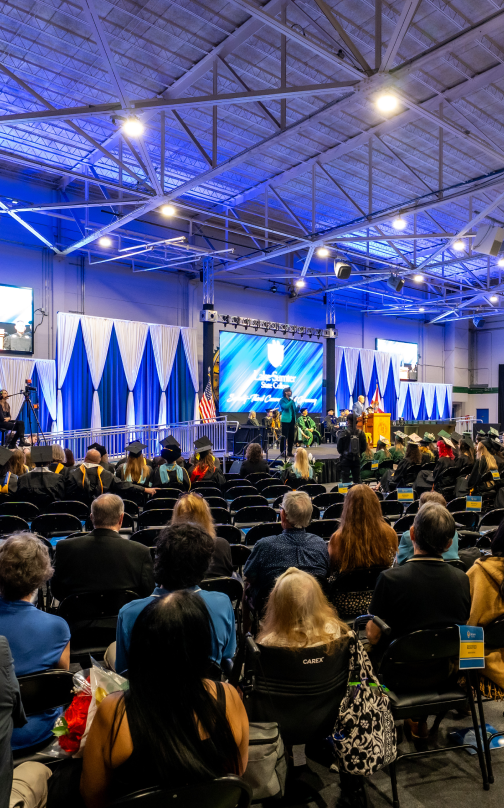 Graduation event, large indoor hall, audience seated, students in graduation attire on stage, blue lighting.